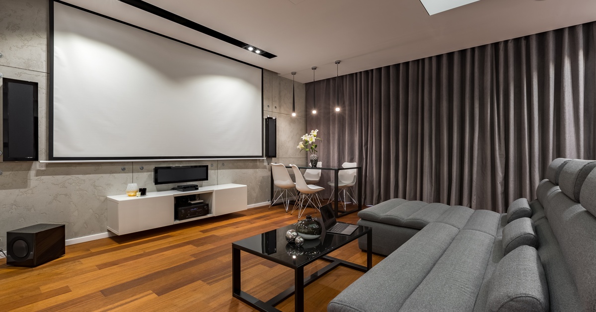 An at-home theater with an L couch in front of a projection screen. The decor palette is black, white, and grey, with wooden flooring.
