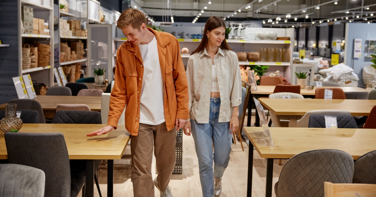 A man and a woman holding hands, walking down an aisle inside a store with multiple desks and chairs on each side.