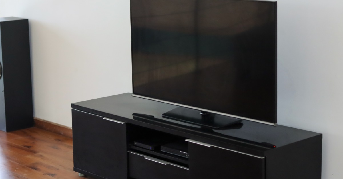 A black entertainment console with silver details holding a large flat screen TV inside a room with wooden floors.