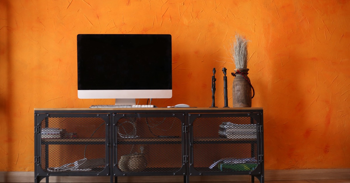 A metal console with wire doors next to an orange wall, holding a large monitor, a plant, and two figurines.