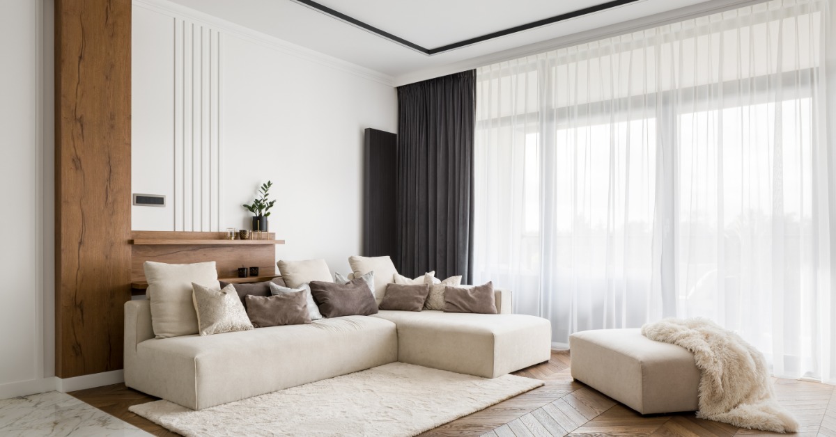 A living room with a white sectional couch over a white area rug, wooden floors, and white sheer curtains.