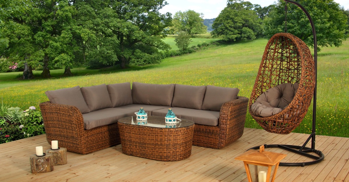 A rattan L-shaped couch next to a hanging swing chair placed on a wooden deck outdoors with a large green field behind.
