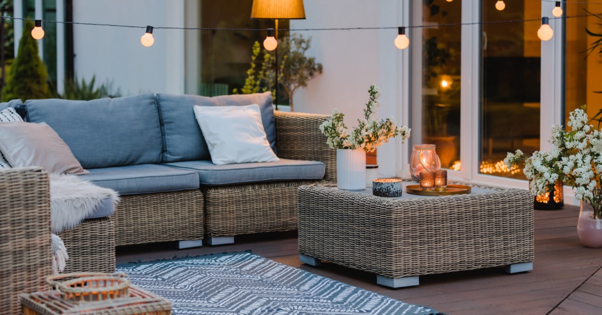 A sectional wicker outdoor couch with blue cushions, a blue rug, light strings, and a lamp next to a large window.