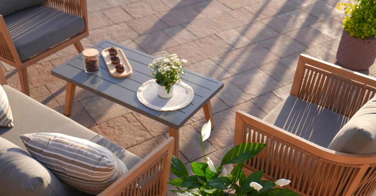 Two wooden chairs and a couch with grey cushions and a coffee table in between, placed on concrete tiles outdoors.