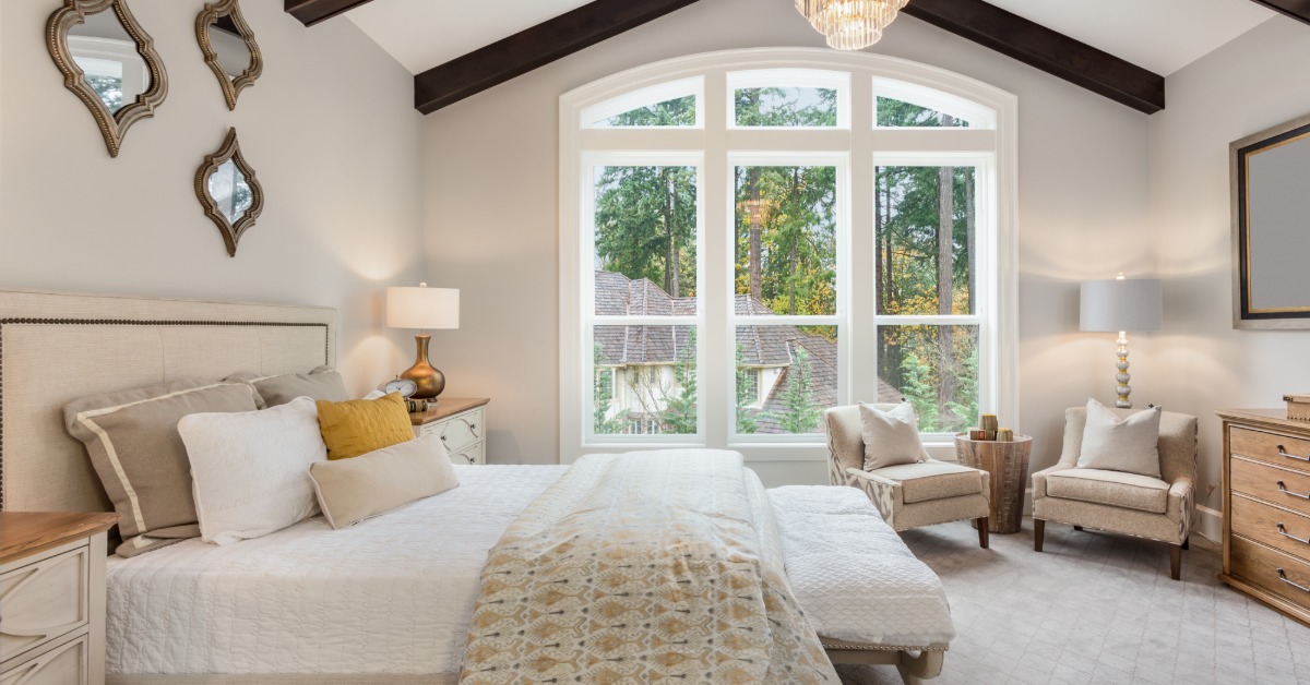 A bedroom with large windows, tiled floor, large cushioned chairs, multiple wall mirrors, and a king-size bed.